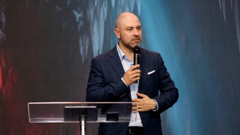 O auto-proclamado profeta Vinicius Iracet, em um púlpito, fala ao microfone, vestindo terno azul e camisa clara. O fundo iluminado e o cenário sugerem que ele está em um evento religioso, fazendo uma pregação