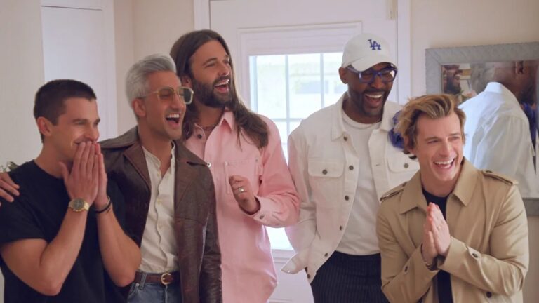 Cinco homens estão lado a lado em um ambiente interno iluminado, sorrindo e rindo com expressões de empolgação e alegria. Um deles usa camisa preta e relógio dourado, outro veste jaqueta marrom com óculos grandes, o terceiro usa camisa rosa, o quarto veste jaqueta branca e boné branco, e o quinto usa sobretudo bege. Ao fundo há uma porta branca com vidro e um espelho refletindo parte da cena.