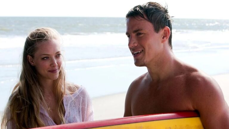 Homem sem camisa segura prancha de surfe e conversa com mulher em praia com mar e ondas ao fundo