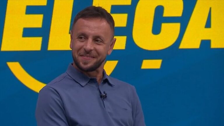 Homem sorri enquanto participa de um programa esportivo, vestindo camisa polo azul, com um painel azul e amarelo ao fundo.