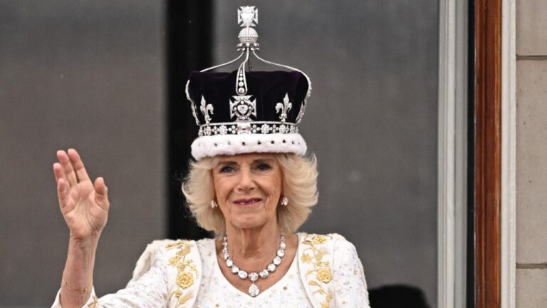 Mulher de cabelos loiros curtos usa manto branco bordado em dourado, colar e brincos de diamantes e uma coroa ornamentada com pedras e detalhes prateados, acenando com a mão levantada diante de uma moldura de janela em cerimônia solene.