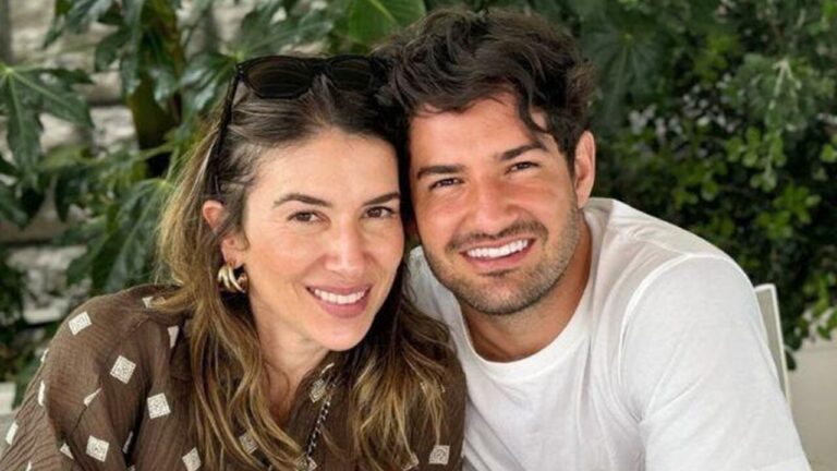 Homem e mulher sorriem juntos em foto ao ar livre, com vegetação verde ao fundo; ele veste camiseta branca e ela usa blusa marrom estampada.