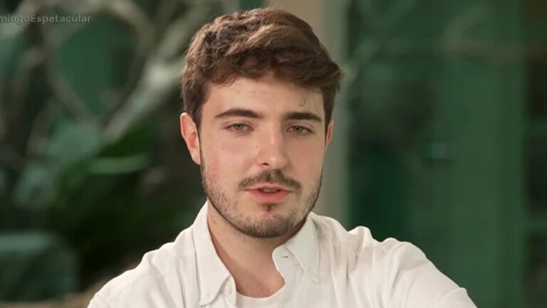 Homem de camisa branca fala durante entrevista em ambiente interno com vegetação ao fundo