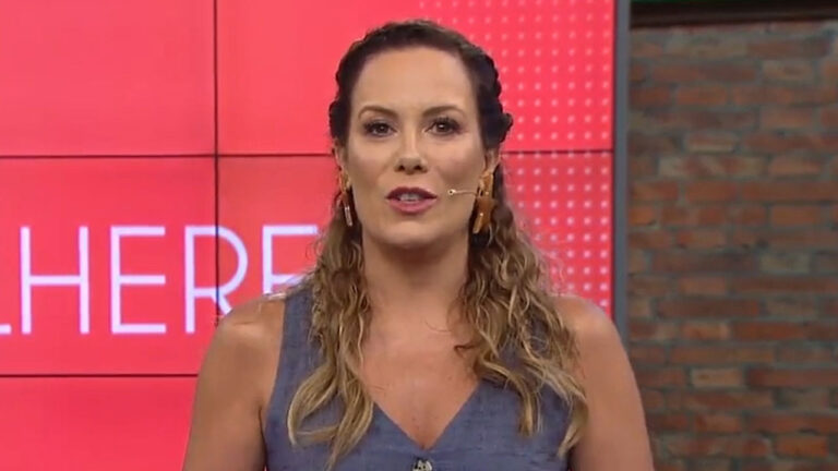 Regina Volpato com blusa azul e microfone de lapela no cenário do programa Mulheres da TV Gazeta diante de telão com logo do programa na cor vermelha