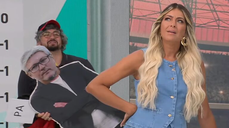 Cena do Jogo Aberto mostra Renata Fan, uma mulher de cabelos loiros longos, usando macacão jeans, com expressão de deboche. Atrás dela, João Paulo Cappellanes, de boné e peruca cinza, segura um recorte de papelão em tamanho real de Odair Hellmann, em um estúdio de TV com temática de futebol