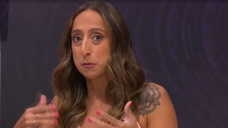 Mulher de cabelos castanhos ondulados e longos fala com expressão séria, gesticulando com as mãos. Ela tem uma tatuagem grande no ombro esquerdo e unhas pintadas de rosa. O fundo é roxo escuro com padrões circulares sutis.