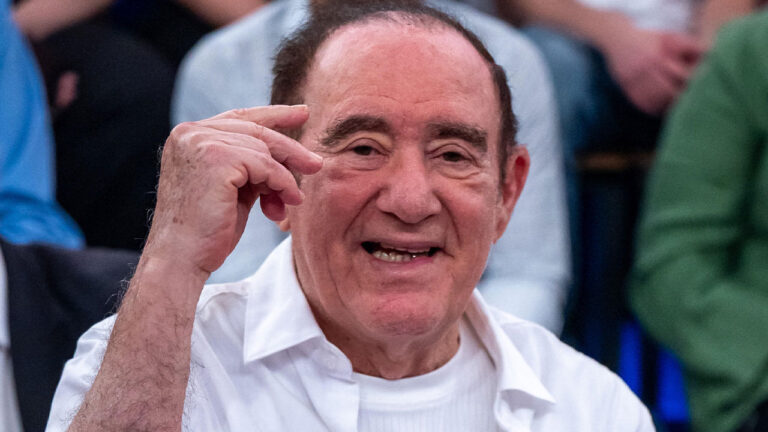 Renato Aragão sorri e acena com a mão próxima ao rosto vestindo camisa social branca durante participação em programa da Globo