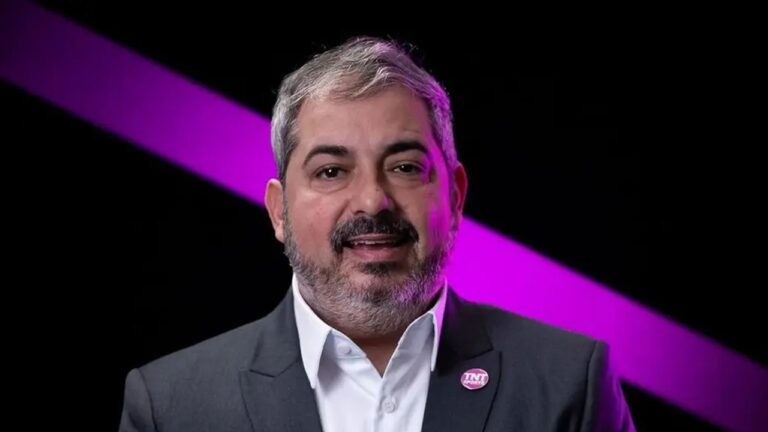 Homem de cabelos curtos grisalhos e barba cheia aparece sorrindo levemente diante de fundo preto iluminado por uma faixa diagonal roxa. Ele veste terno escuro, camisa branca e um broche redondo rosa com logotipo visível preso na lapela.