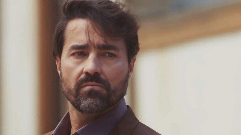 Homem barbudo com expressão séria olha para o lado em cena ao ar livre com fundo desfocado