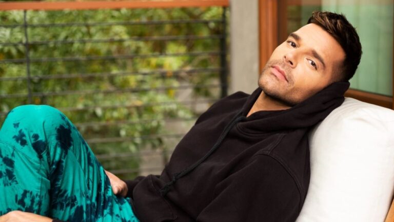 Homem reclinado em um sofá ao ar livre, vestindo moletom preto com capuz e calça verde estampada, olhando para a câmera com expressão relaxada. Ao fundo, há uma varanda com corrimão de metal e árvores verdes desfocadas.