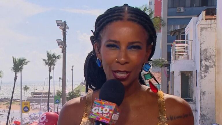 Mulher segura um microfone com o logotipo colorido do “GLO da Rua” e fala diante da câmera em um cenário ensolarado de praia, com o mar, coqueiros e prédios ao fundo. Ela usa vestido dourado brilhante, brincos grandes e coloridos, e maquiagem com sombra azul nos olhos.