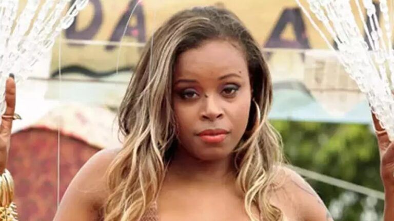 Mulher de expressão séria com cabelo longo e loiro nas pontas, usa argolas grandes e batom vermelho. Ela está ao ar livre, segurando cordas decorativas transparentes com miçangas, e ao fundo há uma placa com letras parcialmente visíveis e árvores desfocadas.