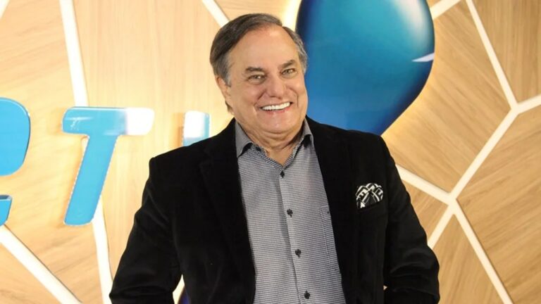 Homem sorri em ambiente interno com cenário de tons amadeirados e azul, vestindo paletó preto, camisa xadrez e lenço no bolso.