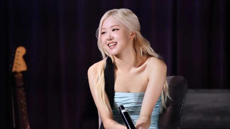 Mulher de cabelos loiros e longos está sentada em uma poltrona, sorrindo e segurando um microfone, com uma cortina escura e um violão ao fundo, sugerindo um ambiente de entrevista ou apresentação musical intimista.