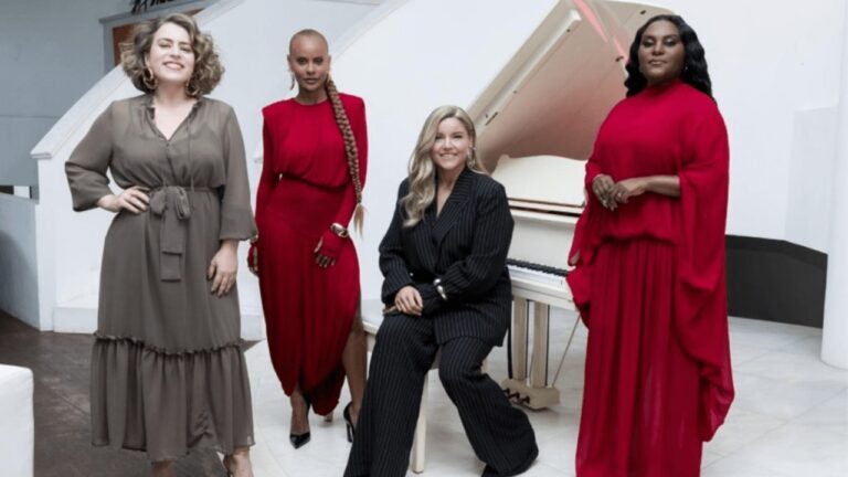 Quatro mulheres posam sorridentes em ambiente elegante, com um piano branco ao fundo; duas usam vestidos vermelhos, uma veste conjunto preto listrado e outra, vestido marrom.