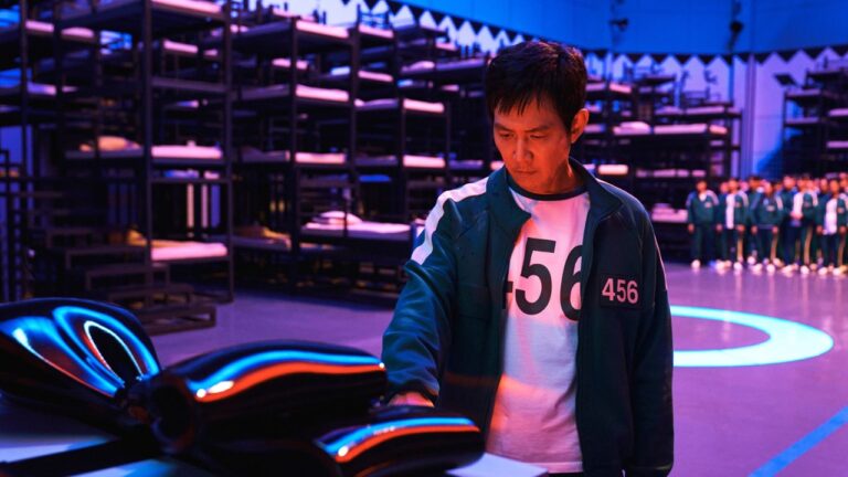 Homem de expressão séria usa um uniforme esportivo verde com o número 456 estampado no peito e toca uma superfície preta brilhante iluminada por luzes coloridas. Atrás dele há várias fileiras de beliches metálicos e um grupo de pessoas vestidas com o mesmo uniforme em pé, em um grande salão com iluminação azul e roxa.