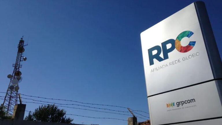 Placa com o logotipo da emissora RPC, afiliada da Rede Globo, e da empresa GRPCOM em destaque; ao fundo, uma torre de transmissão sob céu azul.