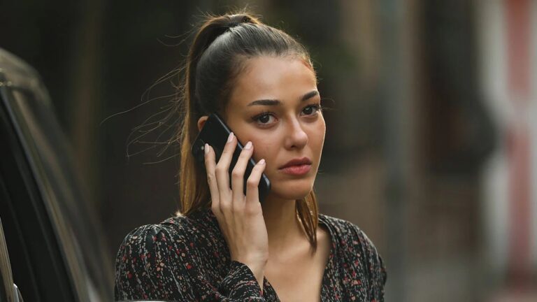 Mulher de expressão séria segura um celular preto próximo ao ouvido enquanto conversa. Ela tem cabelos castanhos presos em rabo de cavalo alto e veste blusa escura estampada com pequenos desenhos coloridos. A cena ocorre ao ar livre, com fundo desfocado mostrando parte de um carro e construções.