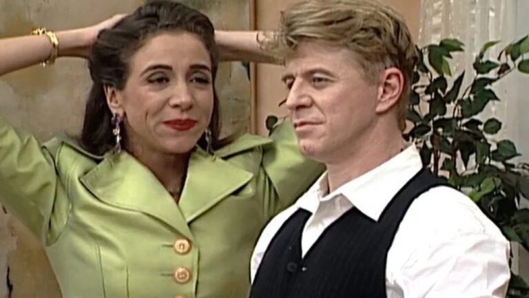 Cena de humor mostra mulher sorridente de blazer verde-claro ao lado de homem loiro de colete preto e camisa branca