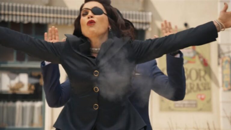 Mulher de tapa-olho abre os braços em cena de ação ao ar livre enquanto é atingida no peito em ambiente urbano