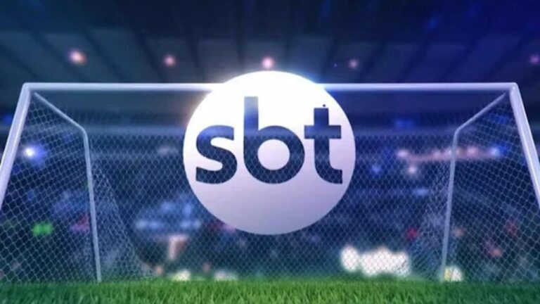 Texto Alternativo disse: Logo do SBT aparece sobre uma trave de futebol em campo iluminado, representando a transmissão esportiva da emissora.