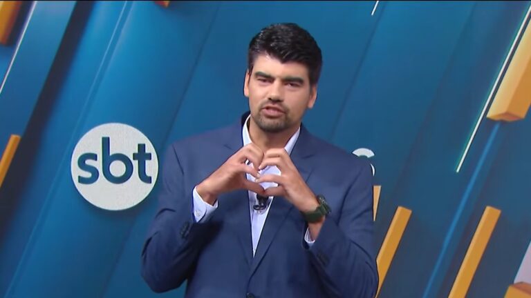 André Azeredo, vestido um terno azul, faz um gesto de coração com as mãos em um estúdio do SBT, cujo logotipo aparece no fundo azul com detalhes em amarelo. Ele apresenta o jornal SBT Notícias 1ª Edição
