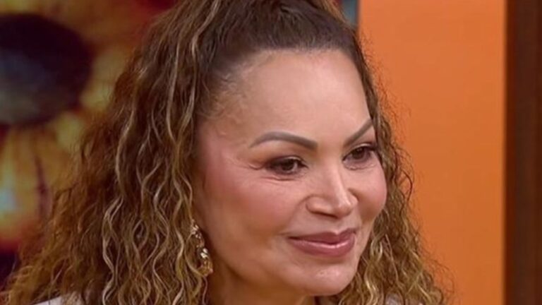 Mulher adulta em close com pele morena clara cabelos longos cacheados em tom castanho claro presos parcialmente no topo da cabeça sobrancelhas bem definidas maquiagem suave com blush rosado e batom nude sorriso discreto e expressão serena usando brincos dourados