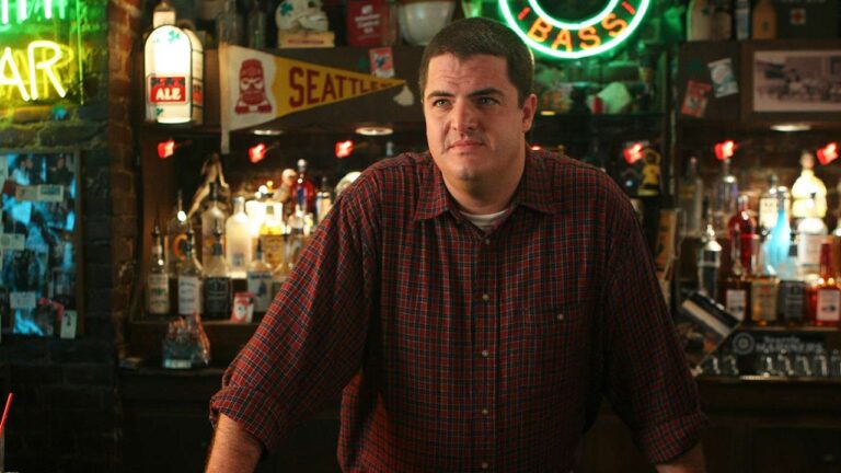 Homem de expressão neutra veste camisa xadrez vermelha e apoia as mãos sobre o balcão de um bar. Ao fundo, há várias garrafas de bebidas, letreiros luminosos e placas decorativas, criando um ambiente típico de bar americano com iluminação colorida.