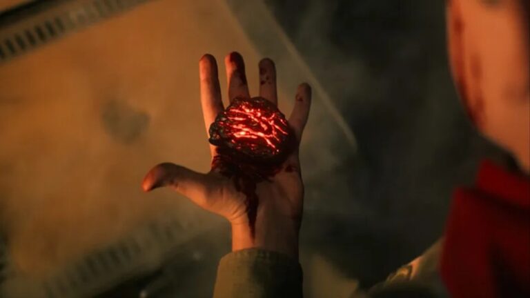 Mão aberta e ensanguentada segura uma pedra escura com rachaduras brilhantes em tom vermelho incandescente, emitindo luz intensa, enquanto o fundo aparece desfocado com tons amarelados e esfumaçados.