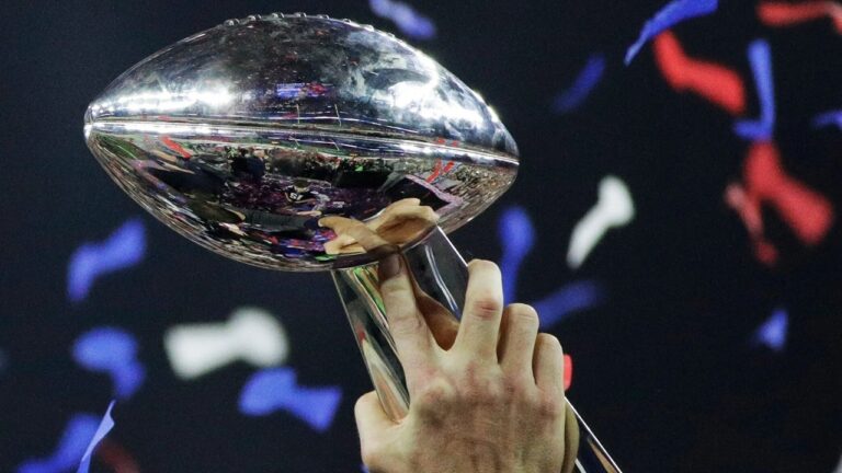 Mão ergue troféu Vince Lombardi refletindo estádio iluminado após final do Super Bowl