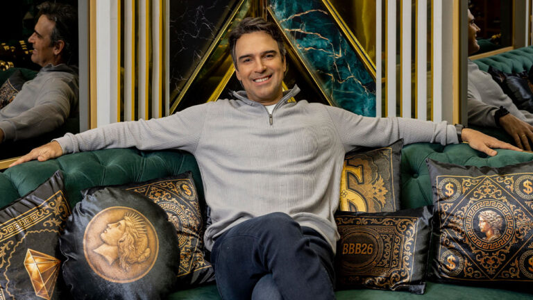 Tadeu Schmidt sorridente sentado em sofá verde com almofadas temáticas do BBB 26 na sala da casa do Big Brother Brasil da TV Globo