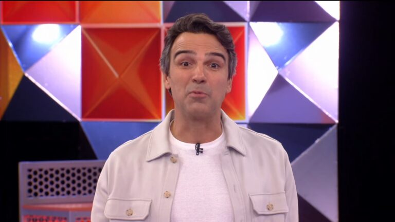 Tadeu Schmidt apresenta a estreia do BBB 26 em estúdio colorido, com painéis geométricos em tons de vermelho, laranja e roxo ao fundo. Ele veste camiseta branca e jaqueta clara, usa microfone preso à gola e tem expressão animada, sugerindo um momento descontraído da atração