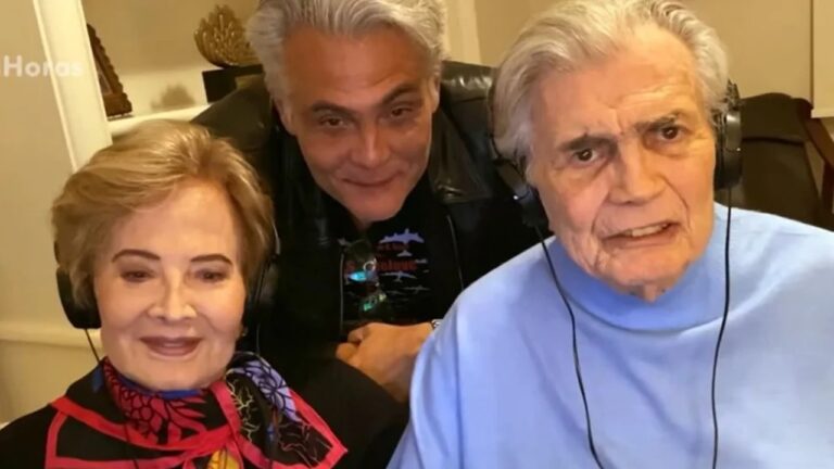 Três pessoas posam juntas em um ambiente interno. Um homem de cabelos grisalhos está ao centro, ligeiramente inclinado para frente, enquanto um casal mais velho, ambos com fones de ouvido, aparece sentado à frente dele. A mulher usa um lenço colorido e sorri, e o homem veste uma blusa azul clara. O clima é de proximidade e carinho, sugerindo uma foto familiar ou de amigos reunidos