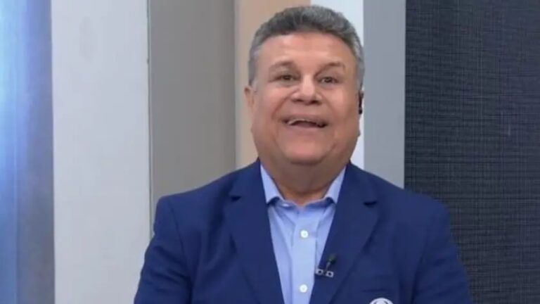 Homem sorri enquanto está em um estúdio de TV, vestindo terno azul e camisa clara, com microfone preso à lapela.