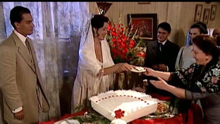 Cena de casamento ambientada em época antiga mostra uma noiva sorridente, vestida com traje branco e véu comprido, entregando um pedaço de bolo para uma convidada idosa que estende as mãos. Um grande bolo branco decorado com flores vermelhas está sobre a mesa coberta por toalha vermelha. À esquerda, o noivo de terno claro observa a cena, enquanto outros convidados elegantes assistem ao fundo em um ambiente interno com cortinas claras e arranjos florais vermelhos.