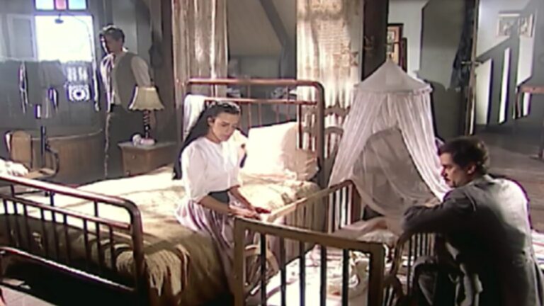 Cena em quarto antigo com iluminação suave mostrando mulher sentada à beira de uma cama de ferro ao lado de um berço coberto por mosquiteiro branco, homem ajoelhado diante do berço e outro homem em pé próximo à janela, ambiente com móveis de madeira e decoração simples.