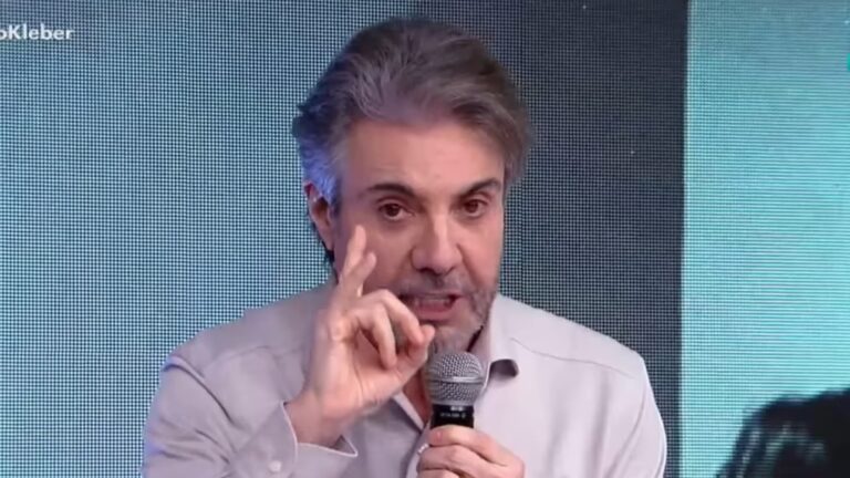 João Kleber, de cabelos grisalhos e barba curta, fala ao microfone enquanto gesticula com a mão, em frente a um fundo iluminado por um grande painel eletrônico. Ele veste camisa social clara e tem expressão séria, apresentando o Teste de Fidelidade na RedeTV!