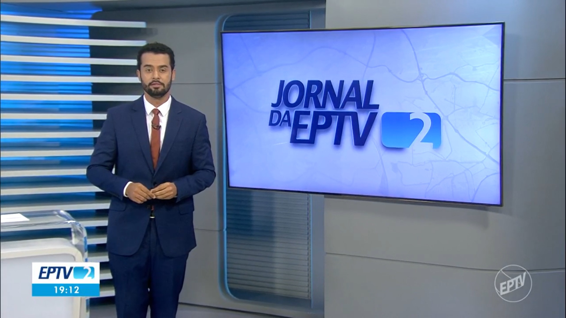 Thales Rodrigues, apresentador do telejornal Jornal da EPTV 2ª Edição, aparece em estúdio moderno com tons de azul e cinza. Ele veste terno azul-escuro e gravata marrom, enquanto o logotipo do programa é exibido em uma tela ao fundo, com o horário 19h12 indicado no canto inferior esquerdo