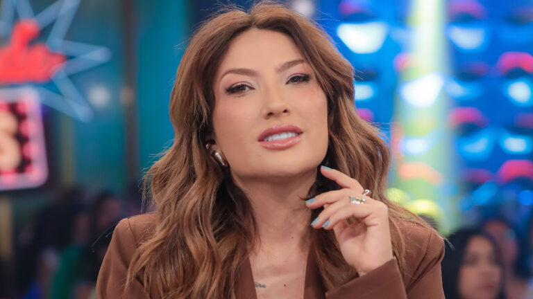 Thayse Teixeira de blazer marrom e unhas azuis posa para foto no palco do Programa Silvio Santos com luzes coloridas e elementos de estúdio ao fundo