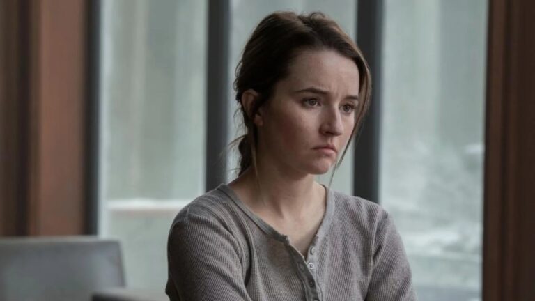 Mulher com expressão triste e olhar distante está sentada em um ambiente interno iluminado por luz natural, vestindo uma blusa simples de manga comprida, transmitindo um clima de melancolia e reflexão.