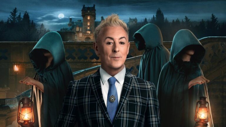 Homem de cabelos loiros curtos e expressão confiante está em primeiro plano vestindo terno xadrez azul e branco, camisa branca e gravata azul com broche. Atrás dele, três figuras encapuzadas em mantos escuros seguram lanternas acesas. O cenário noturno mostra um castelo iluminado ao fundo sob um céu com lua cheia e árvores escuras.