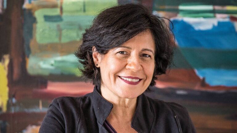 Mulher de cabelos curtos e casaco escuro sorri em frente a pintura colorida em ambiente interno