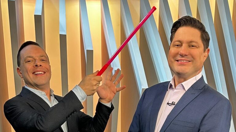 Thiago Gardinali e Eleandro Passaia posam sorridentes no estúdio do Balanço Geral, da Record. Um deles segura uma régua vermelha apontada para o outro de forma descontraída. Ambos vestem ternos e aparecem diante de um fundo com painéis verticais iluminados em tons de bege e azul