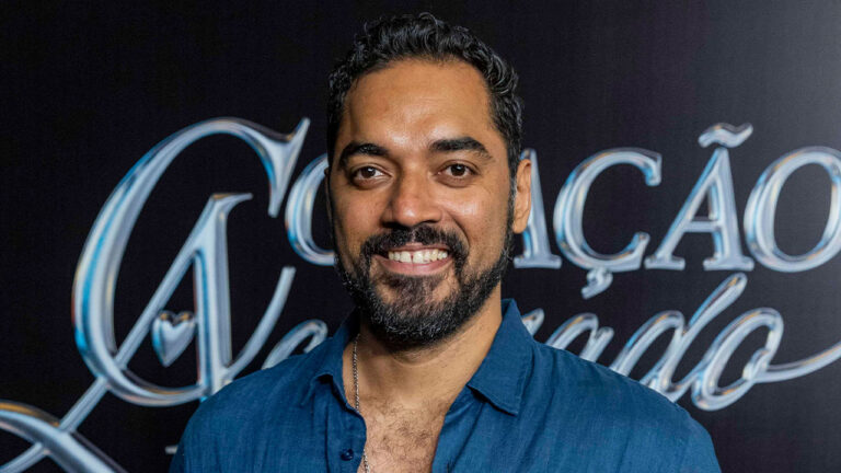 Thomás Aquino sorri com barba e camisa azul em frente ao painel com logo da novela Coração Acelerado