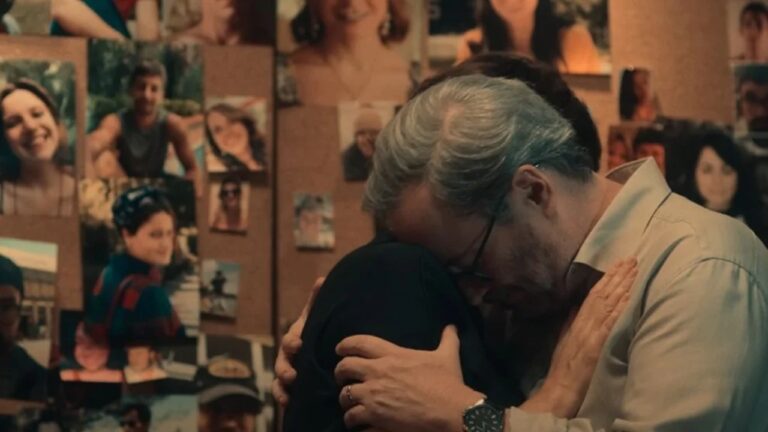 Texto Alternativo disse: Homem de cabelos grisalhos abraça mulher emocionado em frente a mural repleto de fotos em cena de série dramática