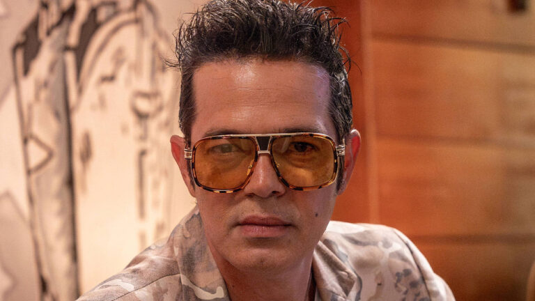 Bruno Mazzeo caracterizado como Tom Bastos usando óculos grandes em cena da novela Coração Acelerado