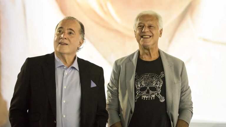 Dois homens estão lado a lado sorrindo em um ambiente interno iluminado. O homem à esquerda usa paletó preto, camisa azul clara e lenço lilás no bolso, enquanto o homem à direita veste blazer cinza e camiseta preta com estampa de caveira feita de brilhos. O fundo é claro e desfocado.