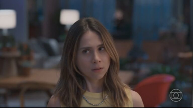 Mulher de expressão séria e olhar pensativo veste blusa amarela sem mangas e colar prateado. O ambiente é interno, com iluminação suave, móveis de madeira e poltronas coloridas desfocadas ao fundo. No canto inferior direito, há o logotipo da TV Globo.