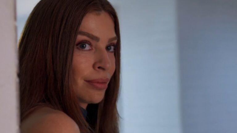 Mulher de cabelos longos e lisos olha por cima do ombro com uma expressão discreta e um leve sorriso. Ela está em um ambiente interno iluminado, com parede branca e fundo desfocado.