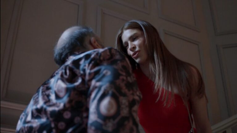 Mulher de vestido vermelho encara de perto um homem de camisa estampada, em cena com iluminação suave e parede de painéis brancos ao fundo, sugerindo tensão entre os personagens.